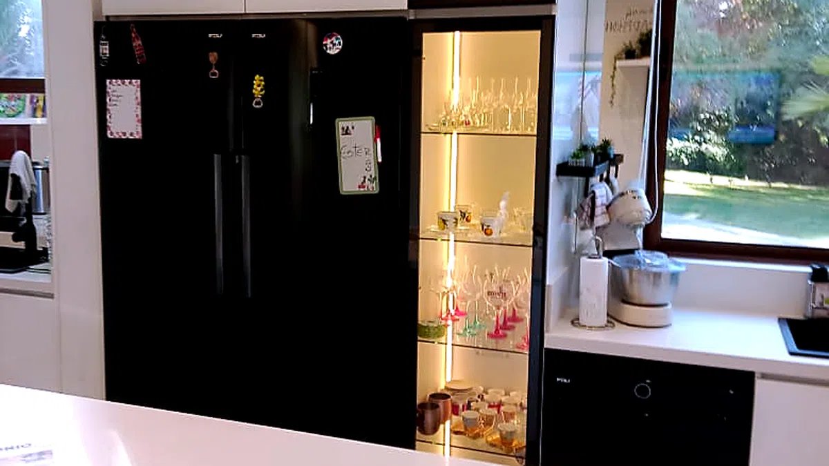 Cocina terminada y en uso: muebles blancos, encimera clara, refrigerador negro de dos puertas y una vitrina iluminada con cristalería, junto a una ventana que da al jardín.